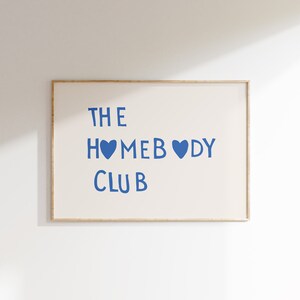 May include: A framed print with the text "THE HOMEBODY CLUB" in blue lettering with two blue hearts. The print is on a white background and is framed in a light wood frame.