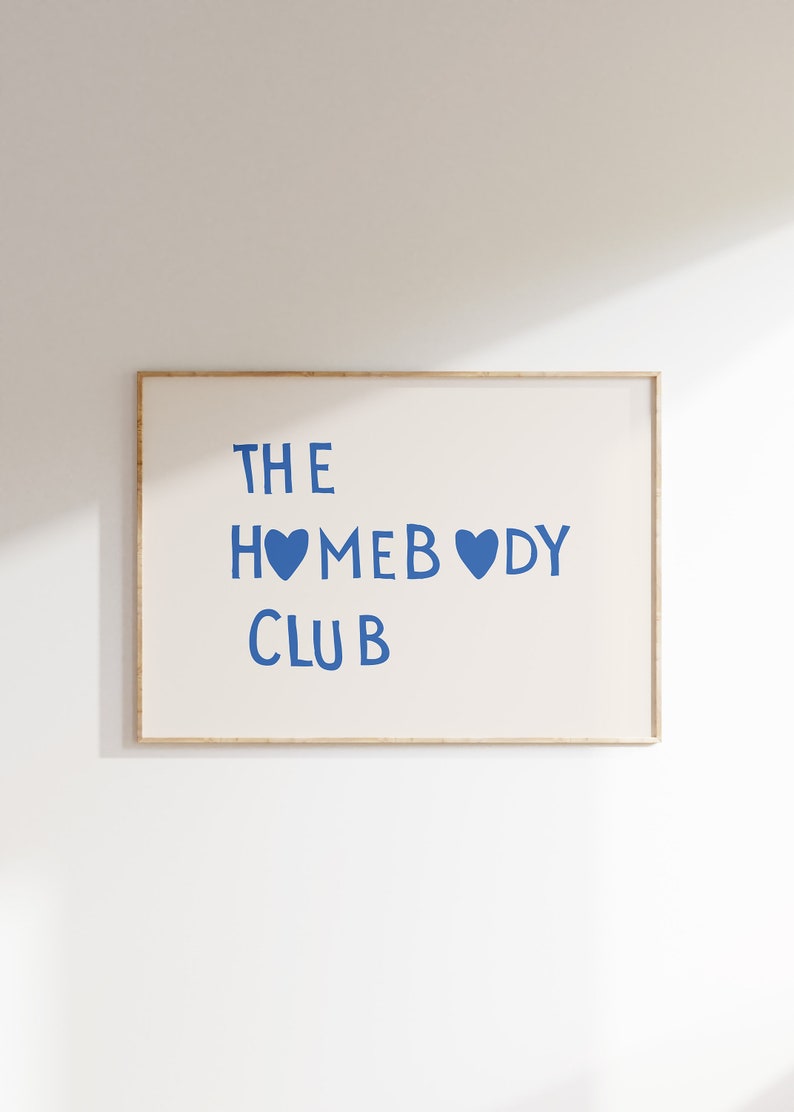 May include: A framed print with the text "THE HOMEBODY CLUB" in blue lettering with two blue hearts. The print is on a white background and is framed in a light wood frame.