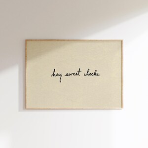 May include: A rectangular framed print with a light beige background. The black cursive text reads "hey sweet cheeks". The frame is a light brown color. The print is hanging on a white wall.