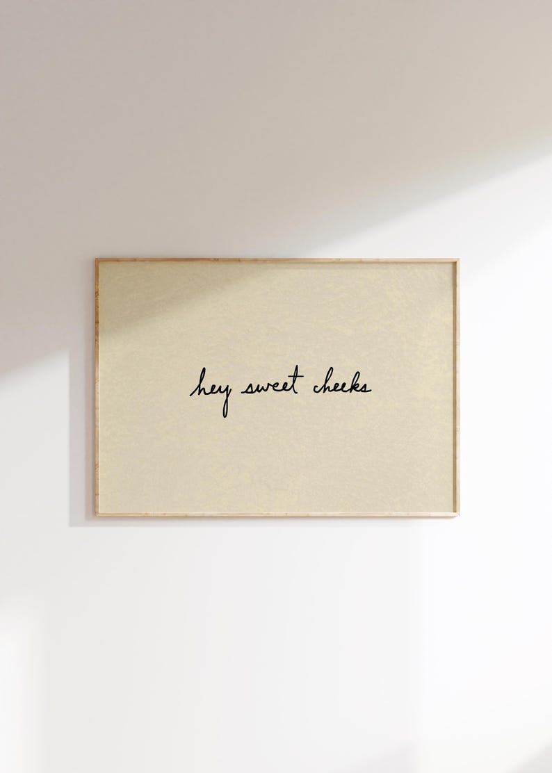 May include: A rectangular framed print with a light beige background. The black cursive text reads "hey sweet cheeks". The frame is a light brown color. The print is hanging on a white wall.