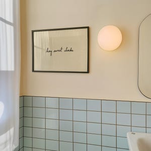 May include: A framed print with the text "hey sweet cheeks" in black script. The frame is black, and the background is a light beige color. The print is hanging on a light beige wall above blue square tiles. A round, white light fixture is to the right.