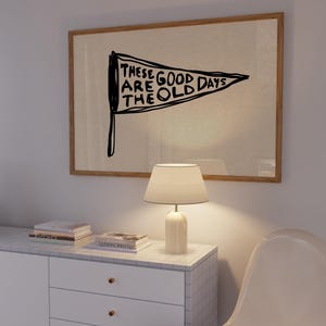 May include: A framed wall art piece with a pennant design. The pennant is black with white text that reads 'THESE ARE GOOD DAYS THE OLD DAYS'. The frame is a light brown color. Below the art is a white cabinet with three drawers and a white lamp with a beige lampshade.
