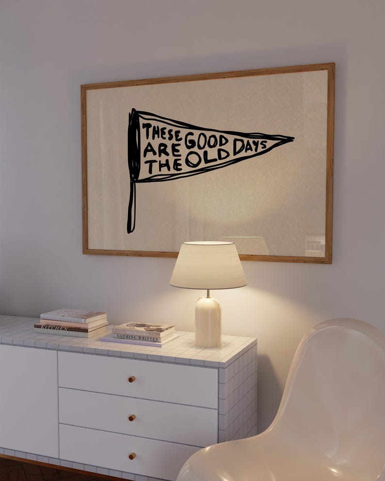 May include: A framed wall art piece with a pennant design. The pennant is black with white text that reads 'THESE ARE GOOD DAYS THE OLD DAYS'. The frame is a light brown color. Below the art is a white cabinet with three drawers and a white lamp with a beige lampshade.