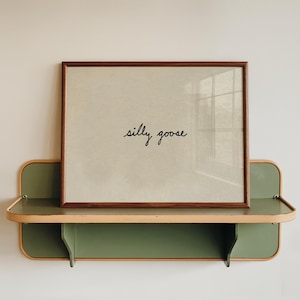 May include: A framed print with the words "silly goose" in black script sits on a vintage-style green and tan metal shelf. The frame is a dark wood color, and the shelf has rounded edges.