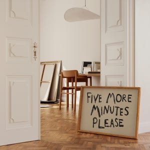 May include: A wooden framed sign with the text "FIVE MORE MINUTES PLEASE" in black handwritten font. The sign is leaning against a wall in a room with a wooden floor and white walls. A wooden chair and desk are visible in the background.