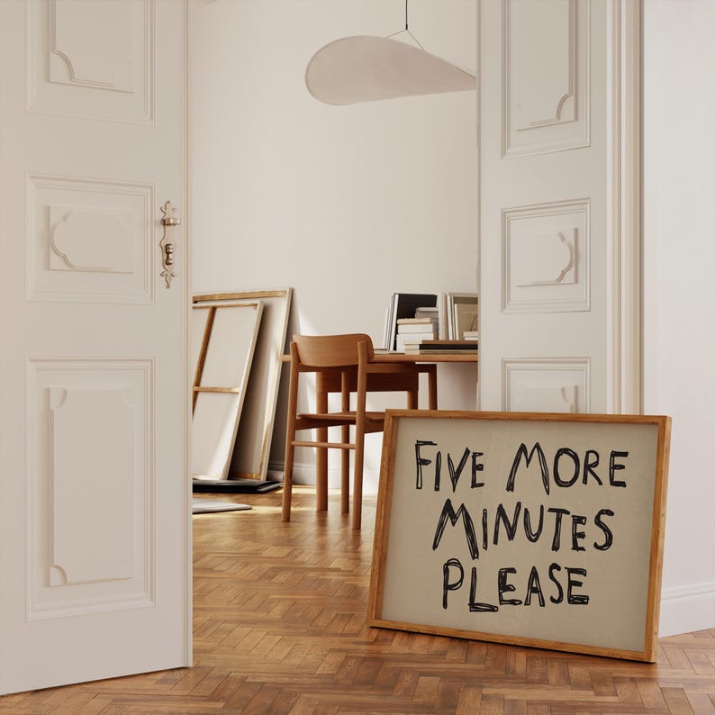 May include: A wooden framed sign with the text "FIVE MORE MINUTES PLEASE" in black handwritten font. The sign is leaning against a wall in a room with a wooden floor and white walls. A wooden chair and desk are visible in the background.