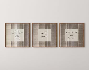 Op de afbeelding: Drie vierkante wandkunstprints in lichte houten lijsten met een verticaal grijs en wit streeppatroon. Elke print bevat een gecentreerd crèmekleurig vierkant met zwarte handgeschreven tekst: THE BEST SEAT IN THE HOUSE, NICE BUM en REMEMBER TO FLUSH.