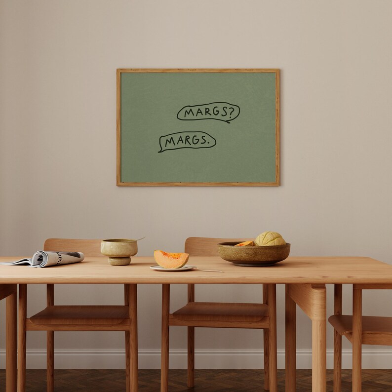 May include: A framed green artwork with the text "MARGS?" and "MARGS." in black speech bubbles. The artwork is displayed above a wooden dining table with chairs, a bowl, and a melon slice.
