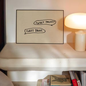 May include: A framed artwork with the text "SWEET TREAT?" and "SWEET TREAT." is displayed on a white surface. A mushroom-shaped lamp and a red book are also on the surface. The scene is illuminated by warm light.