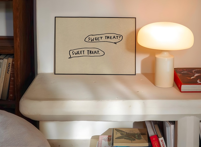 May include: A framed artwork with the text "SWEET TREAT?" and "SWEET TREAT." is displayed on a white surface. A mushroom-shaped lamp and a red book are also on the surface. The scene is illuminated by warm light.