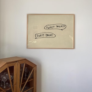 May include: A framed artwork with the text "SWEET TREAT?" above "SWEET TREAT." in black, speech bubble-shaped text. The artwork is set against a light beige background and framed in a light brown wooden frame. The artwork is hanging on a white wall.