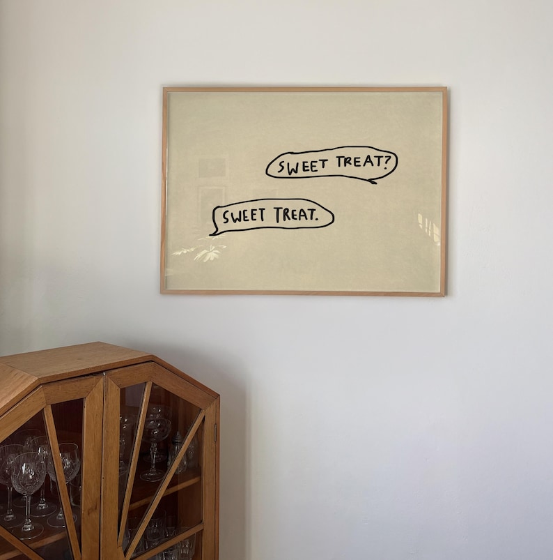 May include: A framed artwork with the text "SWEET TREAT?" above "SWEET TREAT." in black, speech bubble-shaped text. The artwork is set against a light beige background and framed in a light brown wooden frame. The artwork is hanging on a white wall.