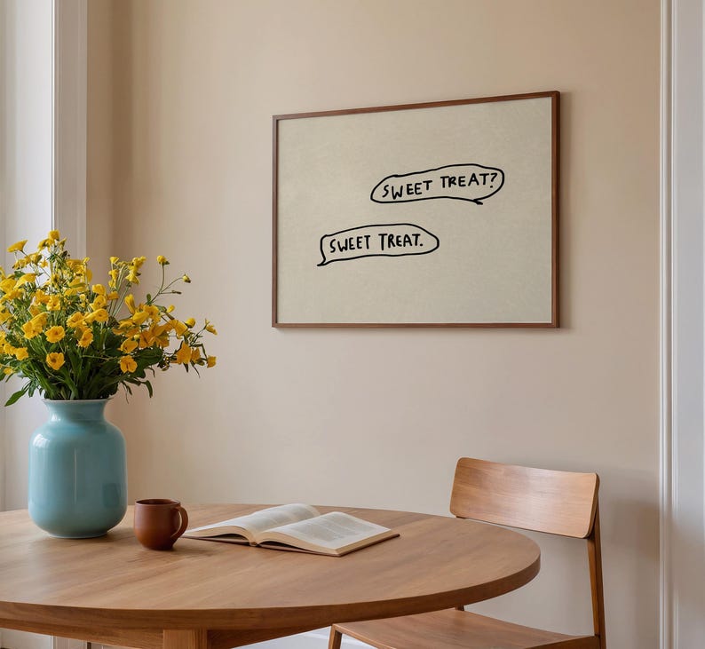 May include: A framed artwork with the text "SWEET TREAT?" and "SWEET TREAT." in black speech bubbles. The artwork hangs above a round wooden table with a vase of yellow flowers and a book. A brown mug sits on the table.