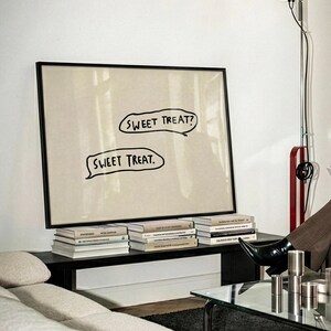May include: A framed artwork with the text "SWEET TREAT?" and "SWEET TREAT." in speech bubbles. The artwork is propped on a dark shelf with several books. A modern floor lamp and a sofa are also visible.