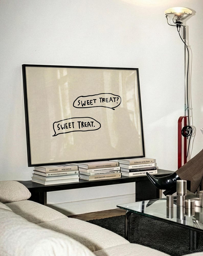 May include: A framed artwork with the text "SWEET TREAT?" and "SWEET TREAT." in speech bubbles. The artwork is propped on a dark shelf with several books. A modern floor lamp and a sofa are also visible.