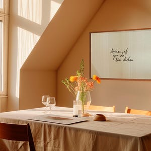 May include: A dining room scene with a wooden table set for a meal. A framed print on the wall reads "kisses if you do the dishes." A vase of flowers and two wine glasses are on the table. The room is bathed in warm sunlight.