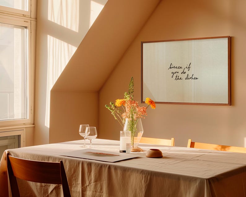 May include: A dining room scene with a wooden table set for a meal. A framed print on the wall reads "kisses if you do the dishes." A vase of flowers and two wine glasses are on the table. The room is bathed in warm sunlight.