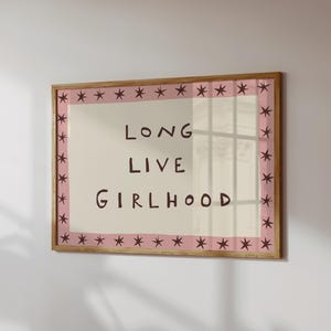 May include: A rectangular wall art print in a wooden frame featuring a cream-colored center with the dark brown text LONG LIVE GIRLHOOD. The border is a soft pink band decorated with a repeating pattern of dark brown eight-pointed stars.