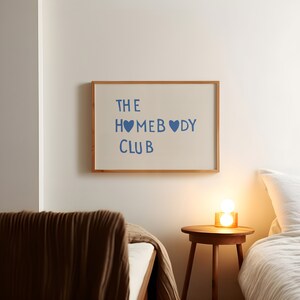 May include: A framed print with the text "THE HOMEBODY CLUB" in blue lettering with two blue heart symbols. The print is hanging on a white wall above a brown bed with a white bedspread and a brown blanket. A small wooden side table with a lamp sits next to the bed.