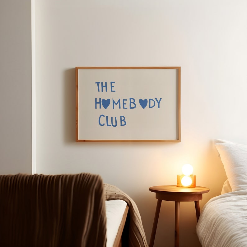 May include: A framed print with the text "THE HOMEBODY CLUB" in blue lettering with two blue heart symbols. The print is hanging on a white wall above a brown bed with a white bedspread and a brown blanket. A small wooden side table with a lamp sits next to the bed.