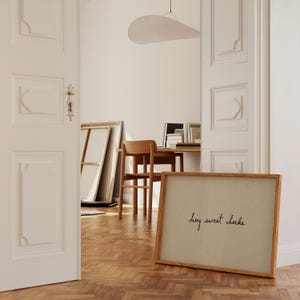 May include: A framed artwork with the phrase "hey sweet cheeks" in black script. The artwork is in a light brown wooden frame and leans against a wall. The room has a wooden floor and a desk with a chair.