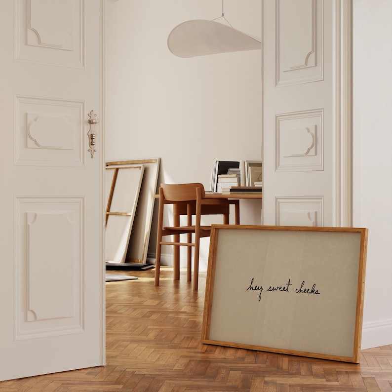 May include: A framed artwork with the phrase "hey sweet cheeks" in black script. The artwork is in a light brown wooden frame and leans against a wall. The room has a wooden floor and a desk with a chair.