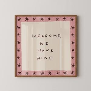 May include: A square framed wall art piece featuring a light beige center with the dark brown text "WELCOME, WE HAVE WINE" in a simple font. The design is bordered by a dusty pink frame decorated with a repeating pattern of dark brown stars.
