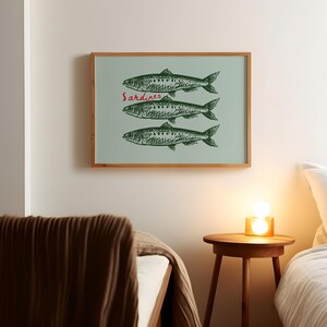 May include: A framed art print featuring three green fish illustrations on a light blue background. The word "Sardines" is written in red script above the fish. The frame is made of wood.