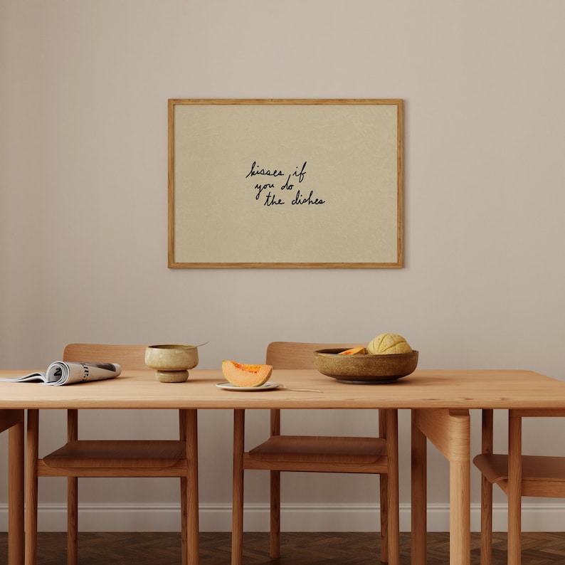 May include: A framed print with the text "kisses if you do the dishes" in a handwritten style. The print is in a wooden frame and hangs above a wooden dining table set with chairs, a bowl, and a piece of cantaloupe.