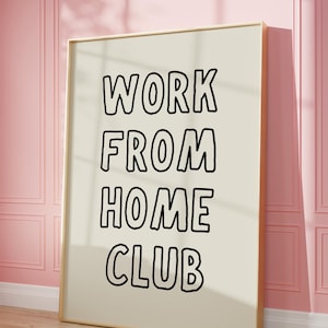 Op de afbeelding: Een ingelijste poster met de woorden "WORK FROM HOME CLUB" in vette, zwarte letters. De poster heeft een cr&egrave;mekleurige achtergrond en een gouden lijst. Hij leunt tegen een roze muur en staat op een houten vloer.