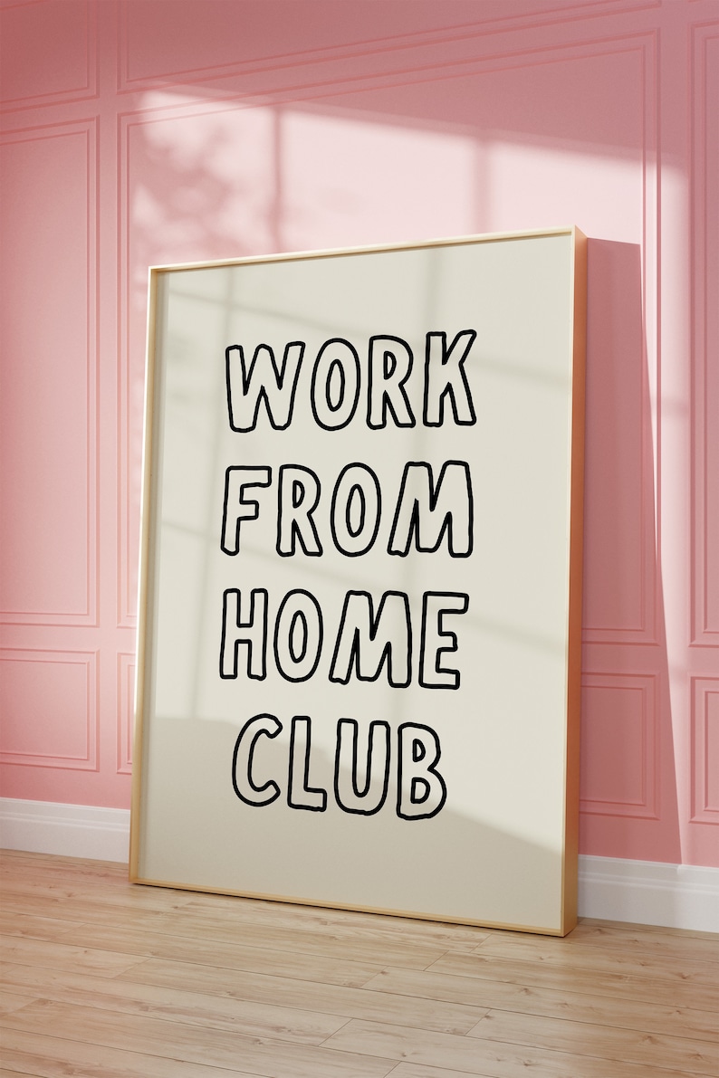 Op de afbeelding: Een ingelijste poster met de woorden "WORK FROM HOME CLUB" in vette, zwarte letters. De poster heeft een cr&egrave;mekleurige achtergrond en een gouden lijst. Hij leunt tegen een roze muur en staat op een houten vloer.