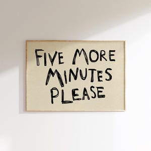 May include: A framed rectangular print with the text "FIVE MORE MINUTES PLEASE" in black handwritten-style font. The background is a light tan color, and the frame is a light brown. The print is displayed against a white wall.