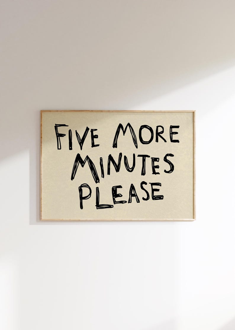 May include: A framed rectangular print with the text "FIVE MORE MINUTES PLEASE" in black handwritten-style font. The background is a light tan color, and the frame is a light brown. The print is displayed against a white wall.