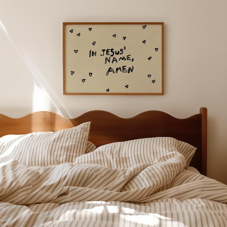 May include: A framed print with the text "IN JESUS' NAME, AMEN" in black, surrounded by small black hearts. The print hangs above a bed with striped bedding and a wooden headboard.