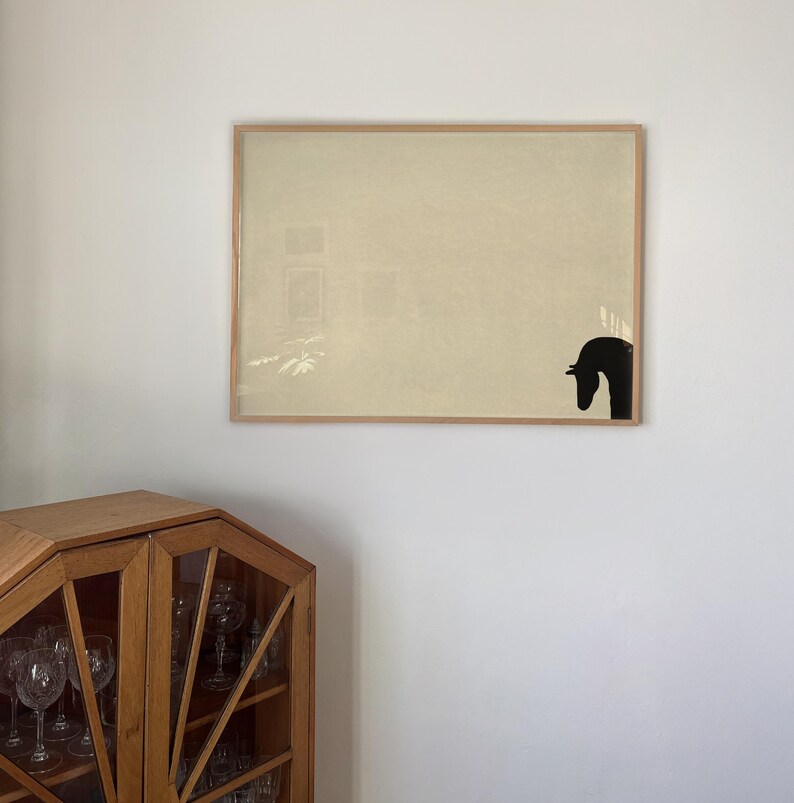 May include: A framed artwork featuring a beige background with a black horse head silhouette in the lower right corner. The artwork is encased in a light brown wooden frame, and is hanging on a white wall.