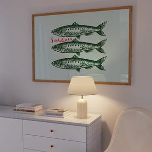 May include: A framed print featuring three green sardines with detailed scales and fins. The word "Sardines" is written in red script above the fish. The print is displayed above a white cabinet with a lamp and books.