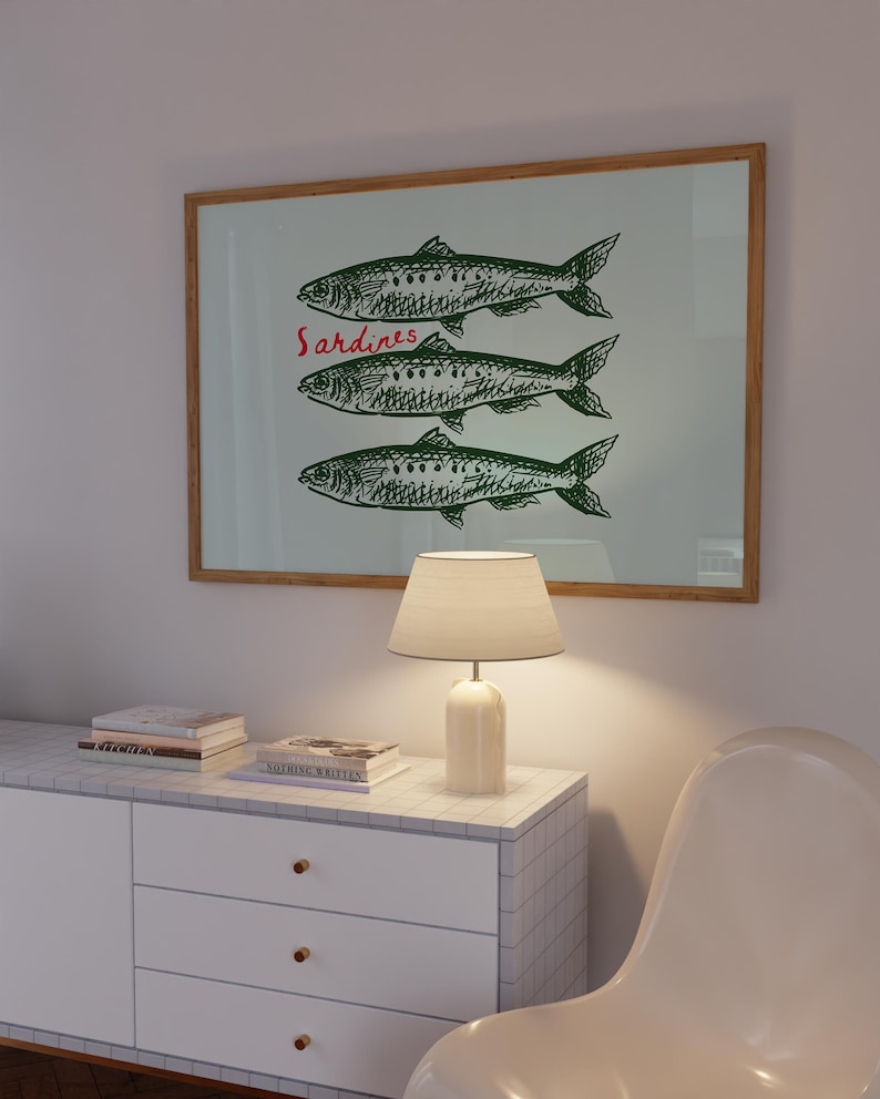 May include: A framed print featuring three green sardines with detailed scales and fins. The word "Sardines" is written in red script above the fish. The print is displayed above a white cabinet with a lamp and books.