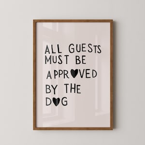 May include: A framed wall art print featuring black text on a light beige background. The text reads ALL GUESTS MUST BE APPROVED BY THE DOG, with black heart shapes replacing the letter O in APPROVED and DOG. The frame is made of medium brown wood.