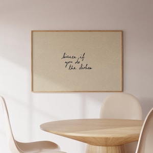 May include: A framed print with the text "kisses if you do the dishes" in black script. The print is mounted on a light beige background and framed in a light brown wood. The print hangs above a wooden table and chairs.