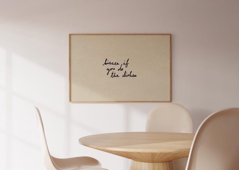 May include: A framed print with the text "kisses if you do the dishes" in black script. The print is mounted on a light beige background and framed in a light brown wood. The print hangs above a wooden table and chairs.