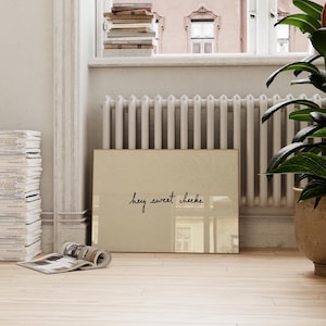 May include: A framed print with the text "hey sweet cheeks" in a cursive font. The print is beige and leans against a white radiator. A stack of books and a potted plant are also visible.