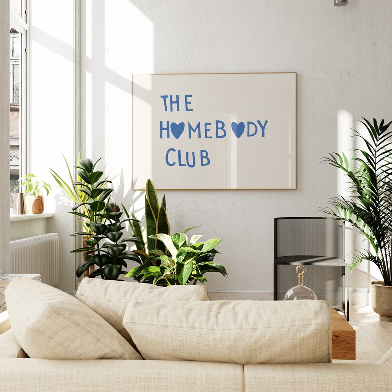 May include: A white framed print with the text "THE HOMEBODY CLUB" in blue lettering with two blue hearts. The print is hanging on a white wall in a living room setting with a couch, plants, and a side table.