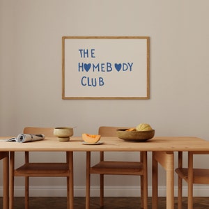 May include: A wooden dining table with three chairs, a bowl of fruit, a plate with a slice of melon, and a ceramic cup. A framed white board with the text "THE HOMEBODY CLUB" hangs on the wall behind the table.