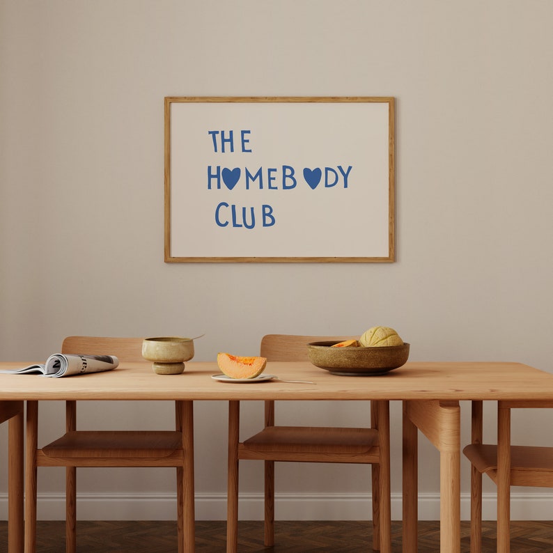 May include: A wooden dining table with three chairs, a bowl of fruit, a plate with a slice of melon, and a ceramic cup. A framed white board with the text "THE HOMEBODY CLUB" hangs on the wall behind the table.