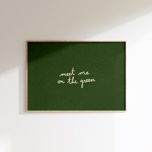 May include: A framed print featuring a dark green backdrop. The words "meet me on the green" are written in cursive script. The frame is a light wood tone, and the artwork is mounted on a white wall, creating a simple aesthetic.