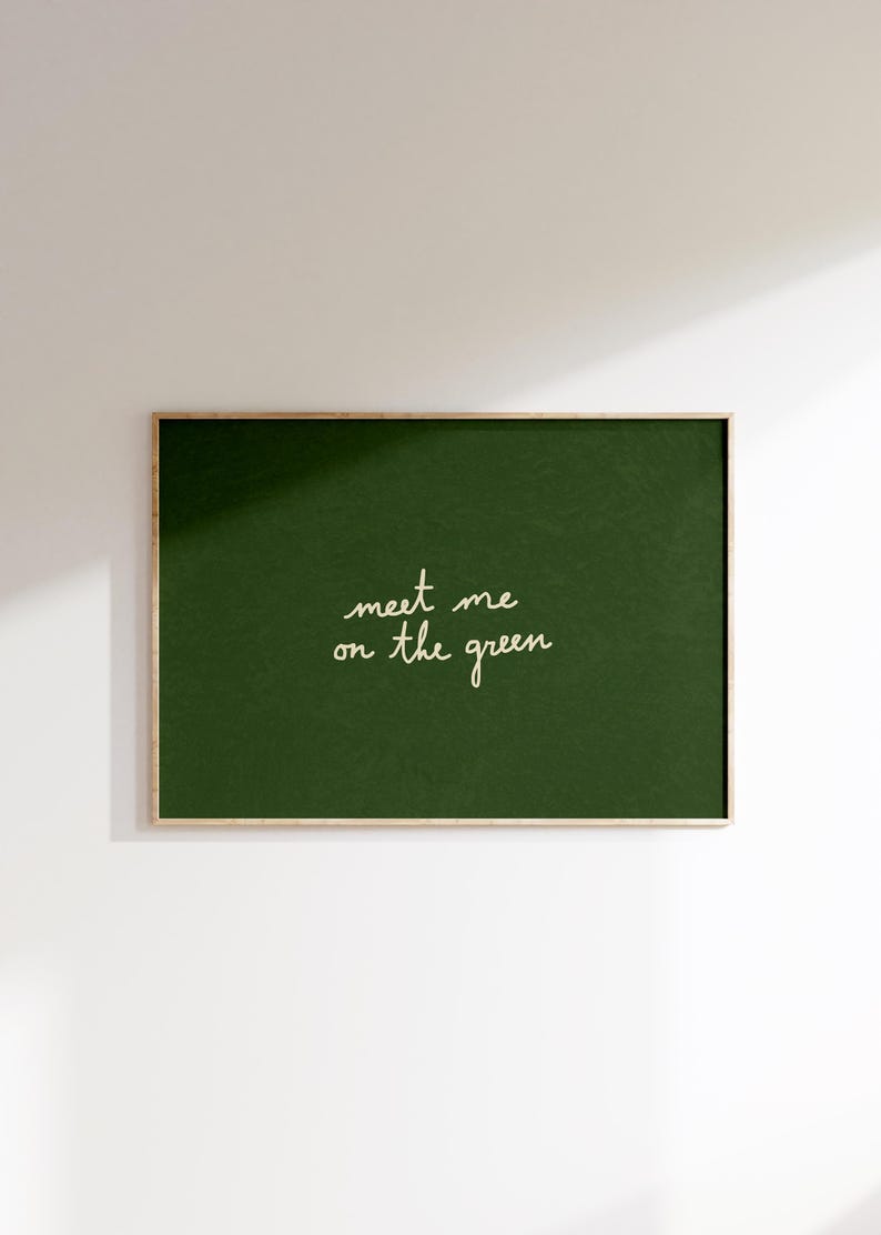 May include: A framed print featuring a dark green backdrop. The words "meet me on the green" are written in cursive script. The frame is a light wood tone, and the artwork is mounted on a white wall, creating a simple aesthetic.
