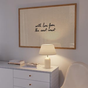 Puede incluir: Una obra de arte enmarcada con el texto "with love from the east coast" cuelga sobre un mueble blanco. Una l&aacute;mpara blanca con una pantalla beige est&aacute; sobre el mueble, junto a libros. Una silla blanca est&aacute; en primer plano.