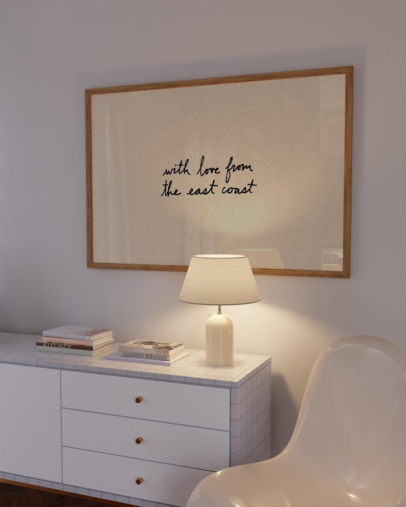 Puede incluir: Una obra de arte enmarcada con el texto "with love from the east coast" cuelga sobre un mueble blanco. Una l&aacute;mpara blanca con una pantalla beige est&aacute; sobre el mueble, junto a libros. Una silla blanca est&aacute; en primer plano.
