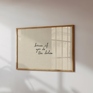 May include: A framed print with the text "kisses if you do the dishes" in black script. The print has a beige background and a light brown wooden frame. The print is hanging on a white wall.