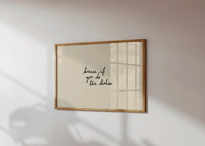 May include: A framed print with the text "kisses if you do the dishes" in black script. The print has a beige background and a light brown wooden frame. The print is hanging on a white wall.